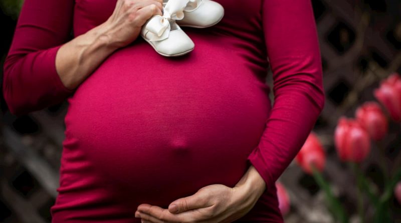 tehotná žena v červených šatách v ruke drží biele detské topánky